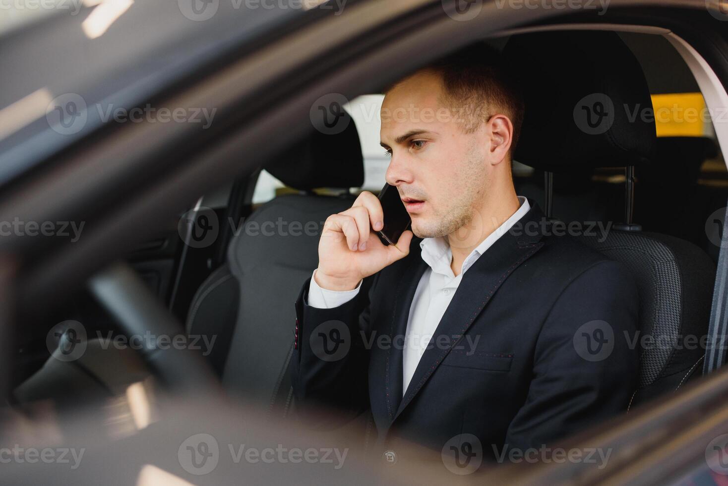 Young businessman sits in luxury car and talks on phone. He looks straight forward. Guy drives car. He holds one hand on steering wheel. It is sunny outside. photo