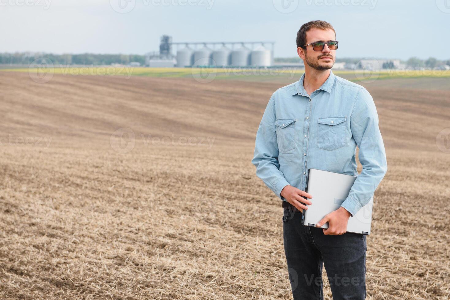 Happy farmer with laptop standing in wheat field in front of grain silo photo