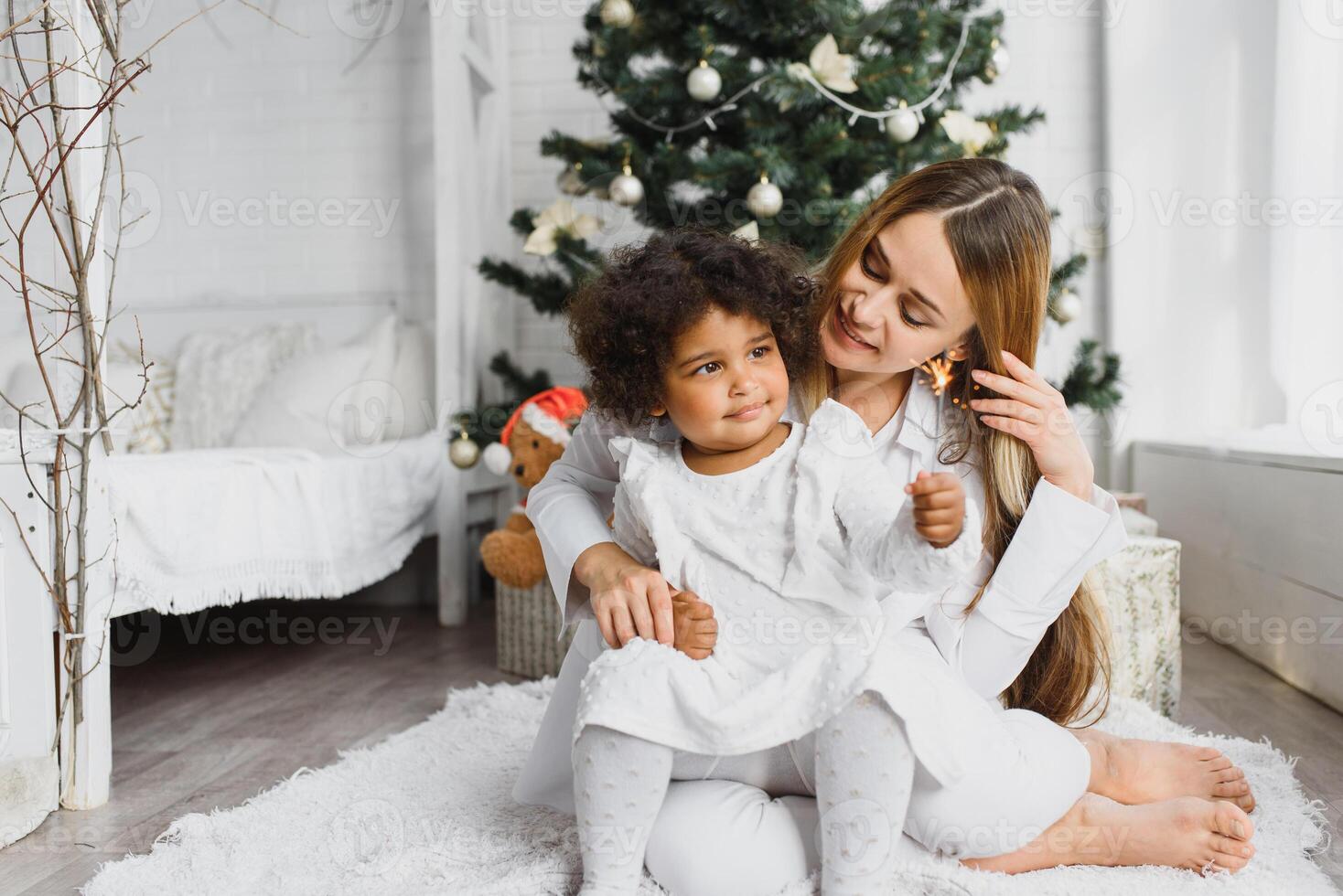 Merry Christmas and Happy Holidays Cheerful mom and her cute daughter at Christmas tree. Parent and little child having fun near Christmas tree indoors. Loving family with presents in room. photo