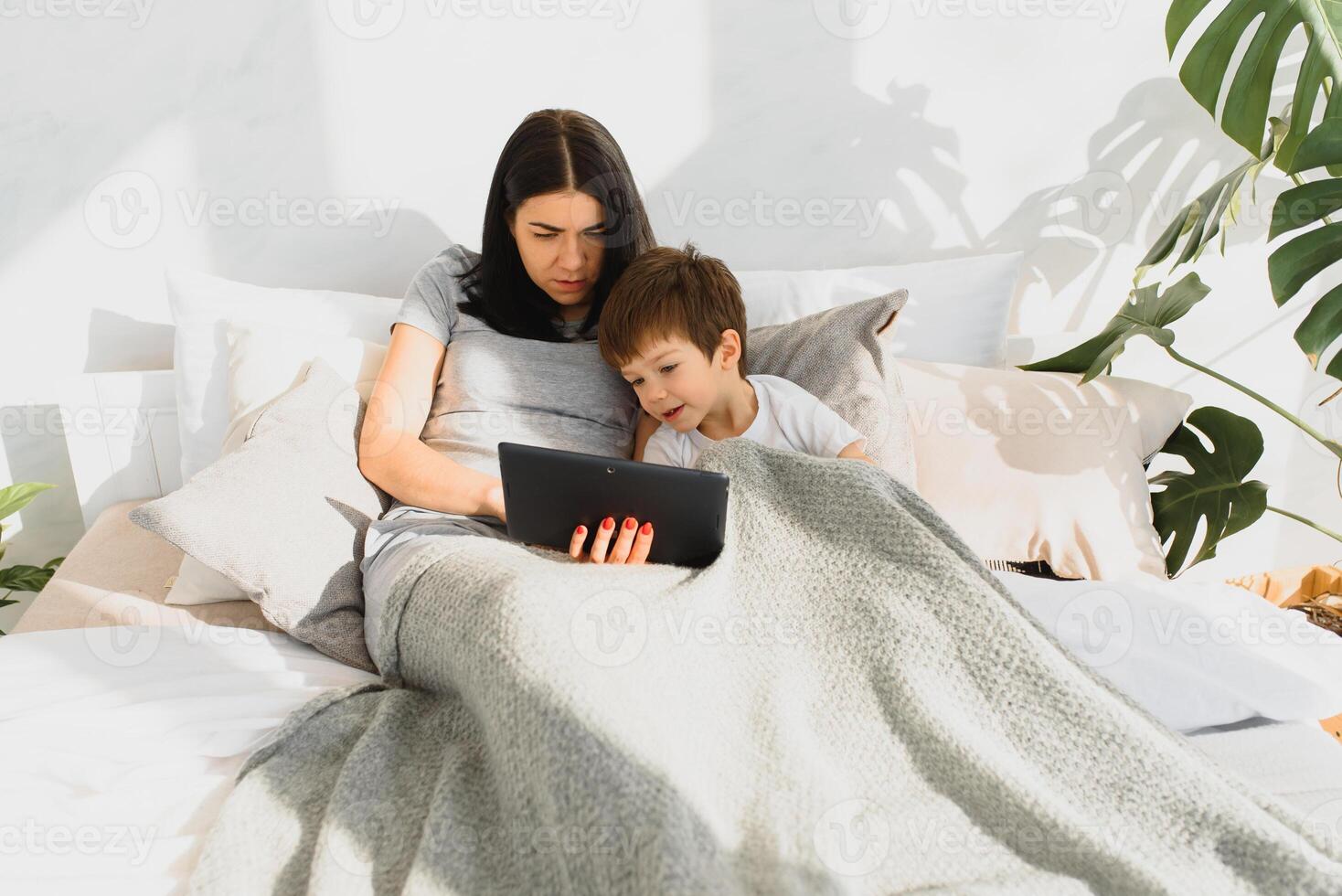 Beautiful smiling mother and son playing with digital tablet laying on bed. photo