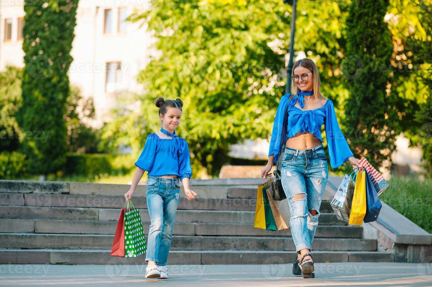 Beautiful mom and her cute little daughter are holding shopping bags, looking at camera and smiling while standing outdoors. Shopping concept. photo