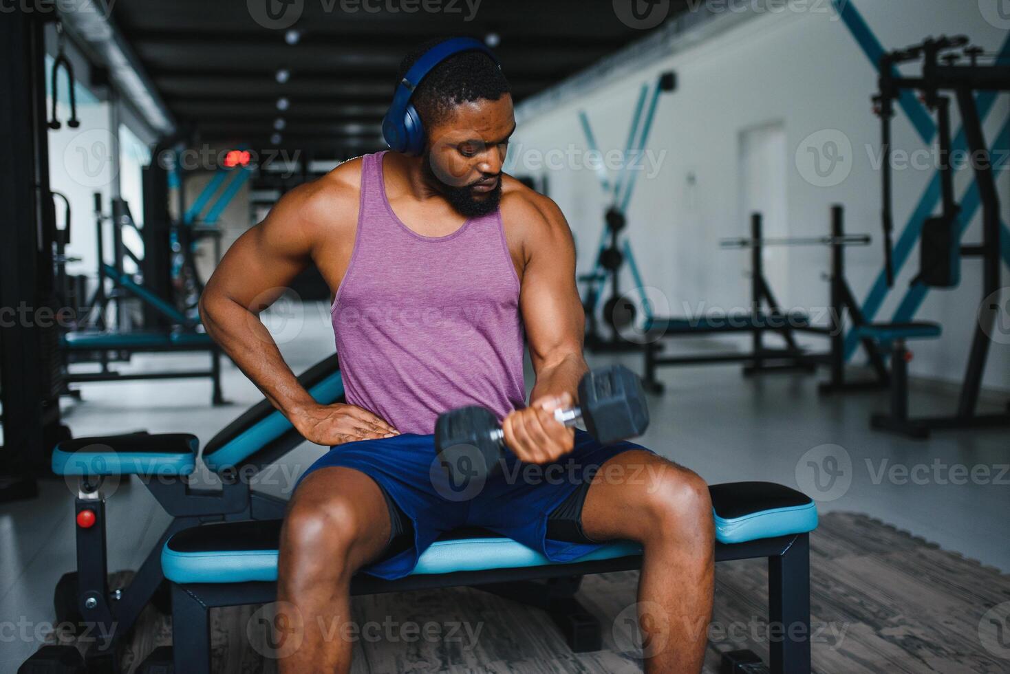 Young African American man sitting and lifting a dumbbell close to the rack at gym. Male weight training person doing a biceps curl in fitness center. photo