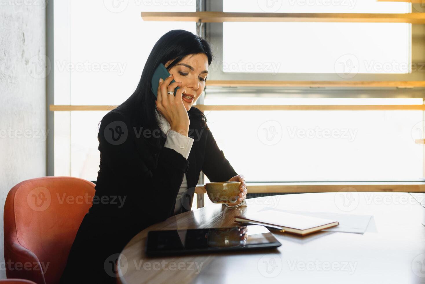 Cheerful young business woman standing in modern office talking on her mobile phone. Beautiful female model using cell phone. photo