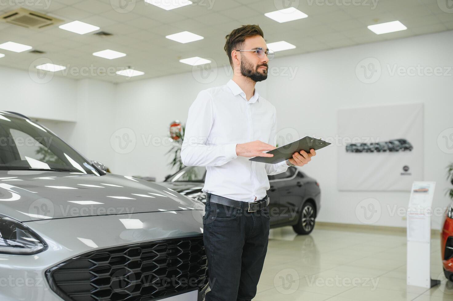 de cerca Disparo de exitoso confidente sonriente caucásico masculino tienda asistente participación portapapeles en formal ropa a automóvil coche comerciante tienda foto