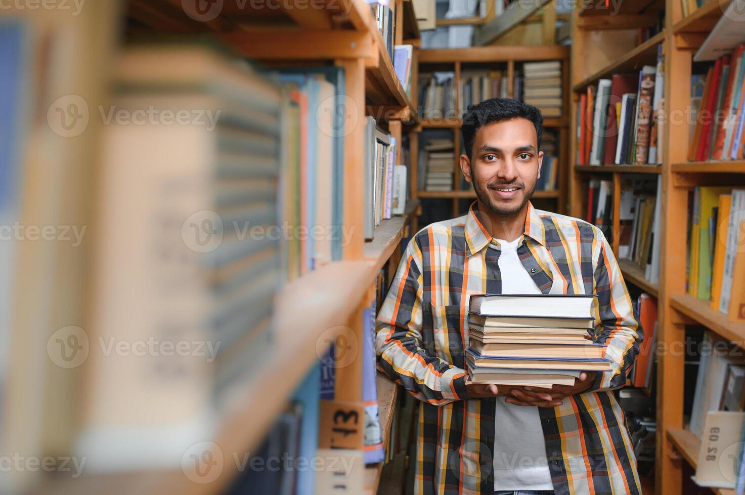 Portrait of cheerful male international Indian student with backpack, learning accessories standing near bookshelves at university library or book store during break between lessons. Education concept photo
