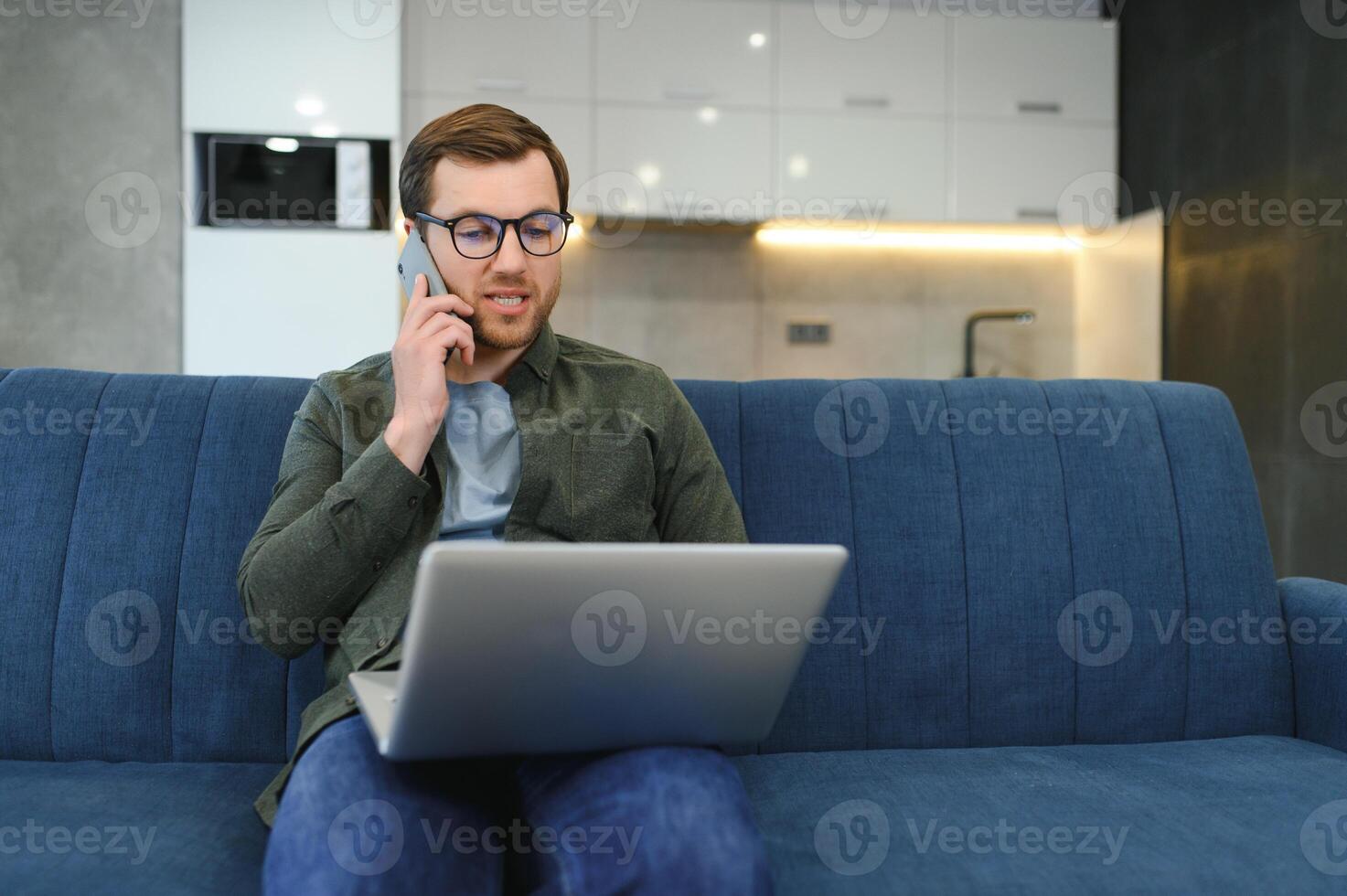 Cheerful busy young caucasian male manager sits on sofa with computer, calls by phone, talks with client, looks at free space in white room interior. Business, work remotely at home, ad and offer. photo