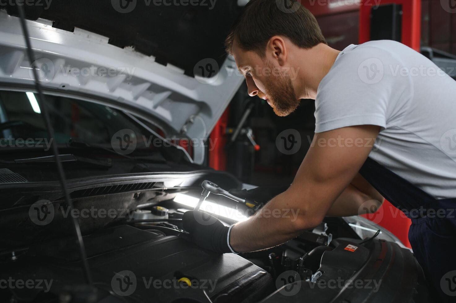 hermoso joven masculino auto mecánico en especial uniforme ropa participación un Linterna, mirando para Descompostura y reparando debajo el capucha en el coche motor en un coche taller foto