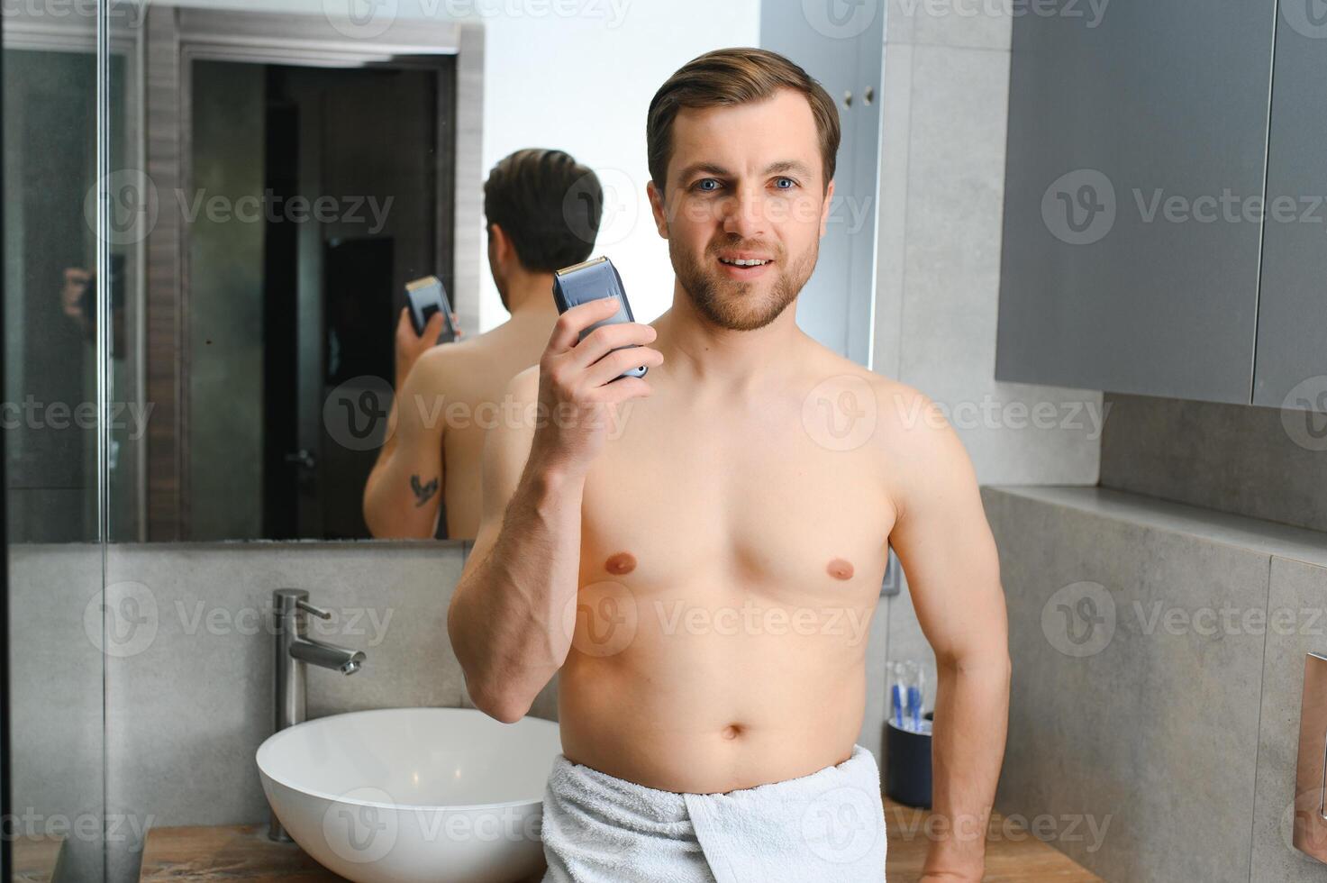 beauty, shaving, grooming and people concept - close up of young man looking to mirror and shaving beard with trimmer or electric shaver at home bathroom photo