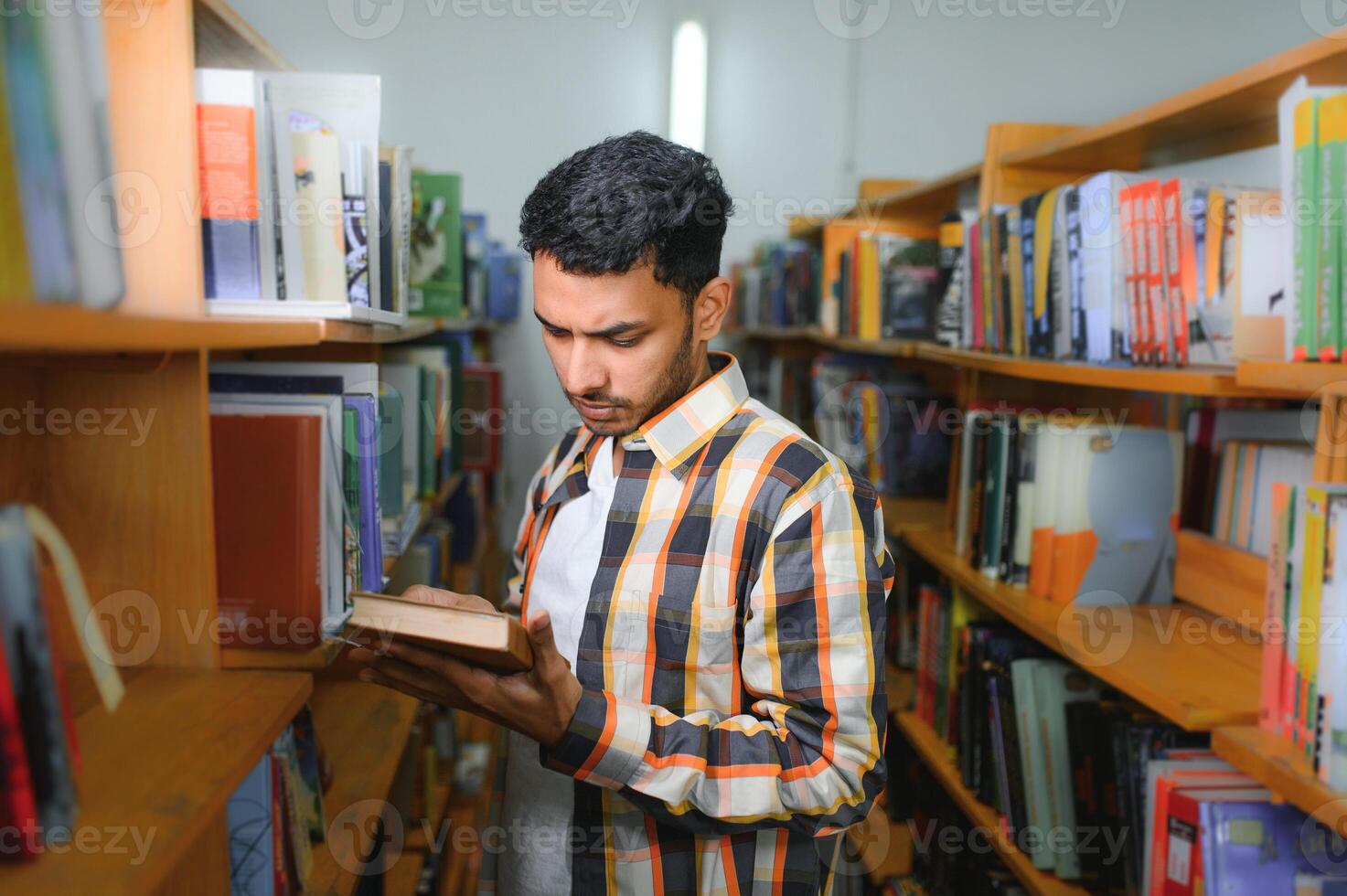 Portrait of cheerful male international Indian student with backpack, learning accessories standing near bookshelves at university library or book store during break between lessons. Education concept photo