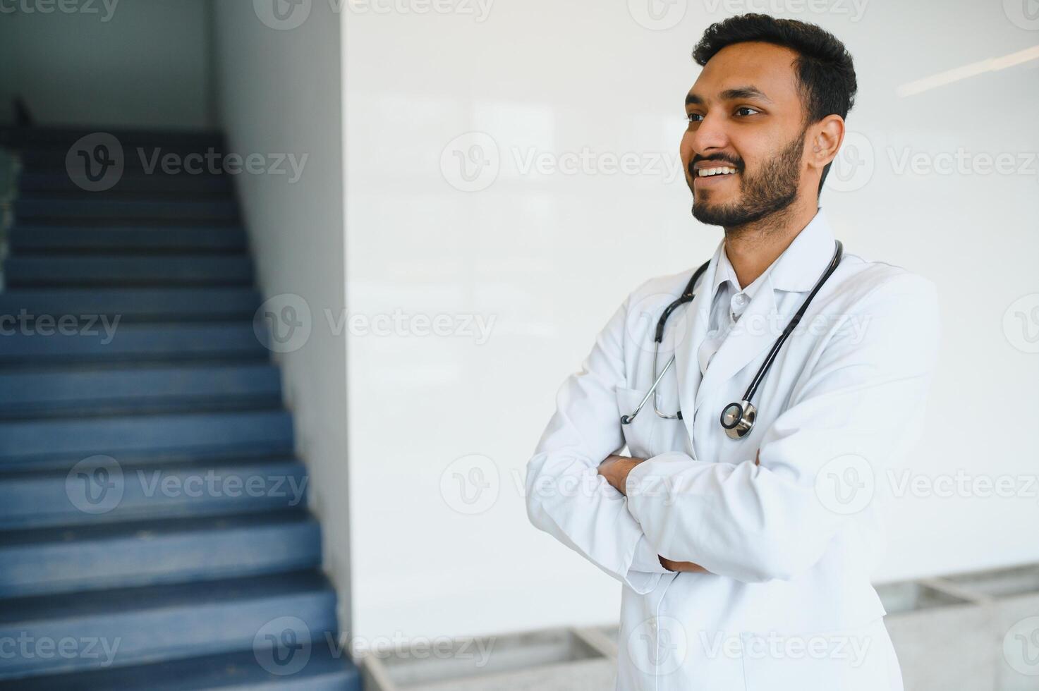 Portrait of male indian doctor wearing white coat having open door on clinic corridor as background photo