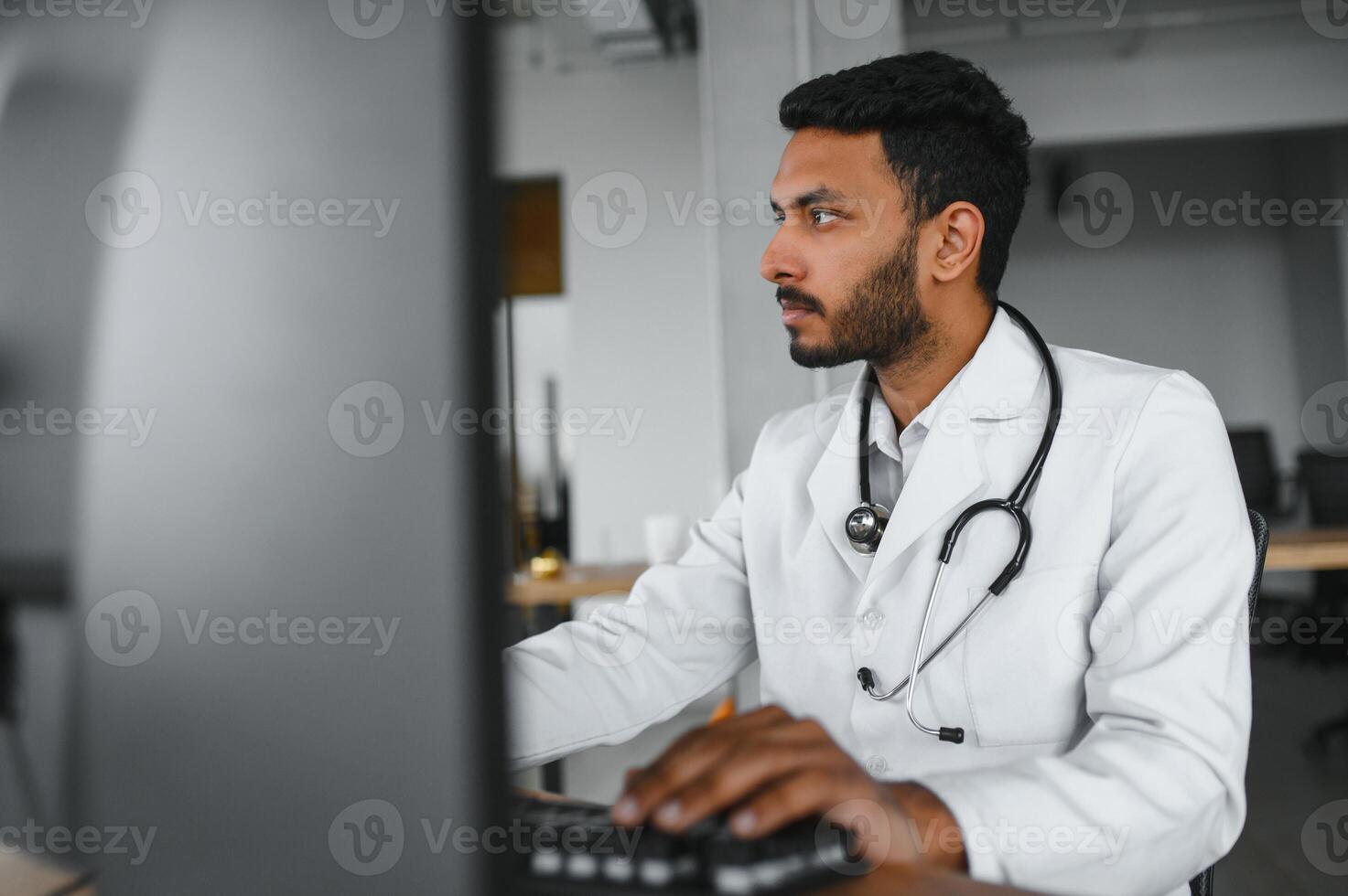 People, occupation and medicine concept. Smiling male Indian Arab doctor in white coat, sitting in medical office at desk with laptop photo