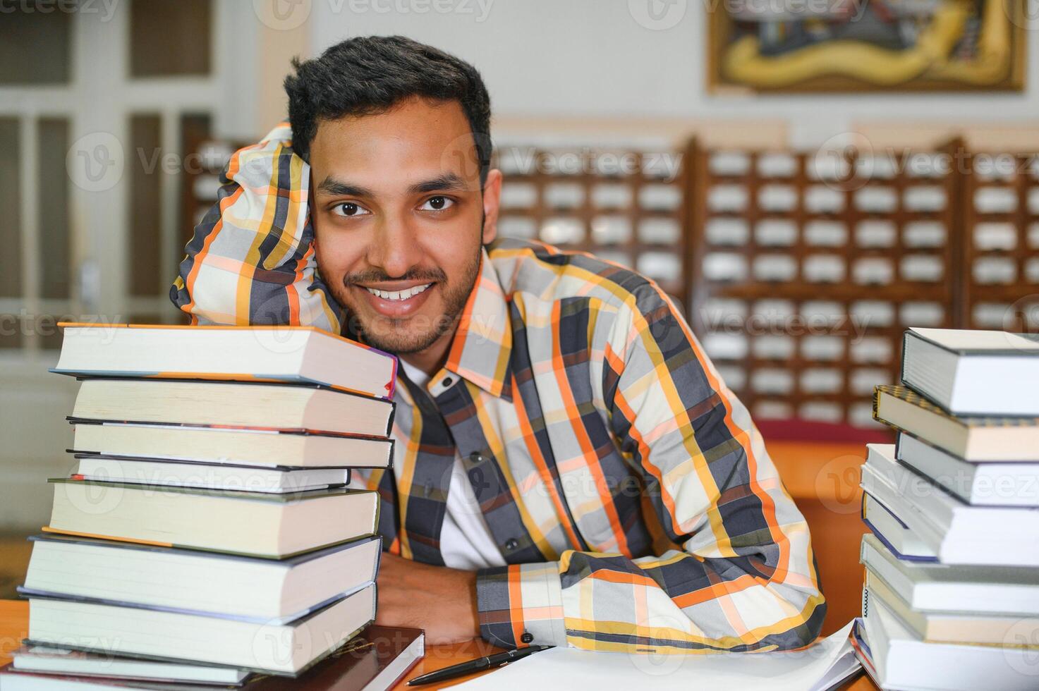 Portrait of cheerful male international Indian student with backpack, learning accessories standing near bookshelves at university library or book store during break between lessons. Education concept photo