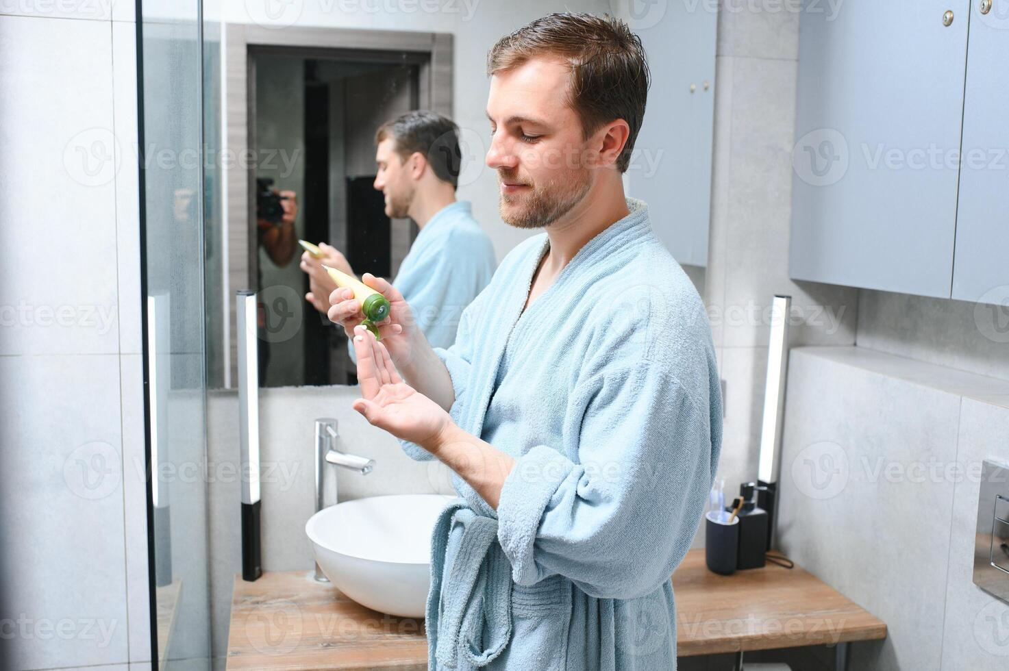 Handsome young man applying cream on his face in bathroom photo