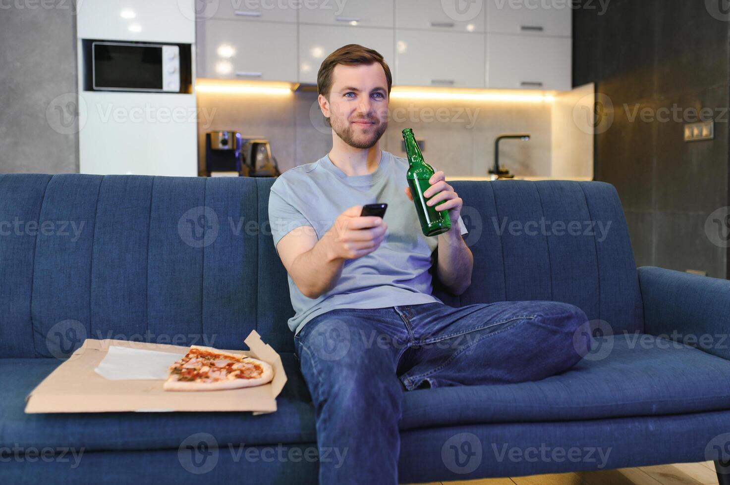 Eats pizza while watching TV show. Man with beard indoors. photo