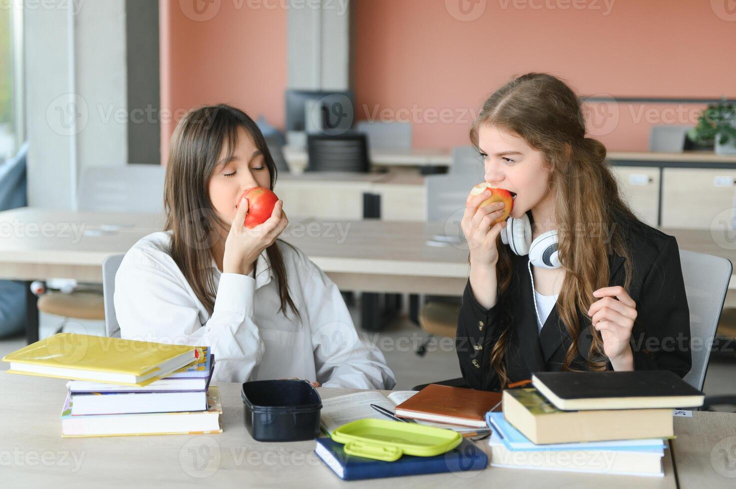 un linda niña sonriente a su compañera de clases mientras teniendo almuerzo a el escritorio en colegio durante un descanso foto