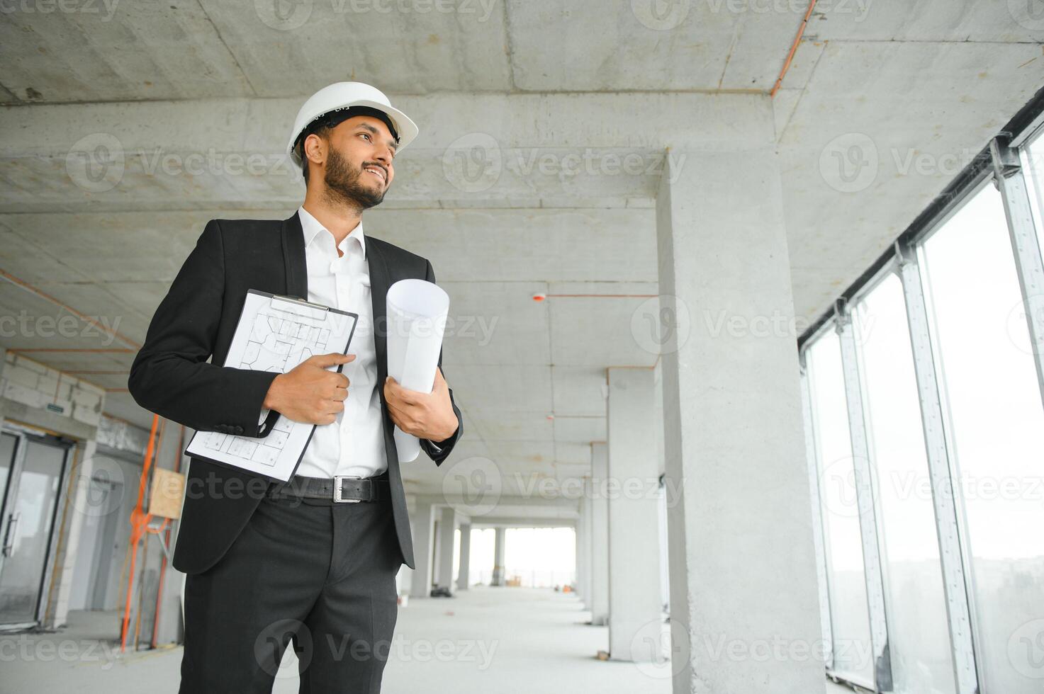 Indian construction site manager standing wearing helmet, thinking at construction site. Portrait of mixed race manual worker or architect. photo