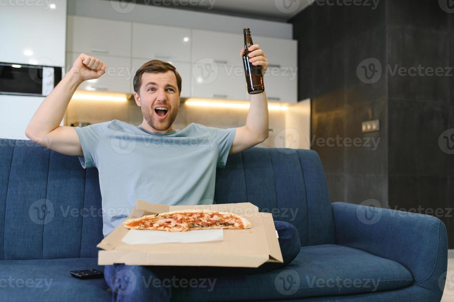 Happy young man drinking beer and eating pizza when watching game on tv at home photo