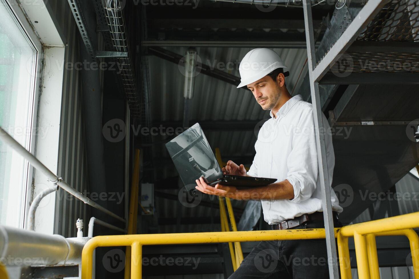 A young handsome employee of a modern factory in a white hard. In the background is a large shop for the production. photo