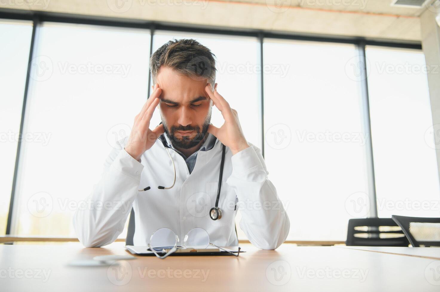 Handsome doctor man wearing medical uniform sitting on his workplace tired holding his head feeling fatigue and headache. Stress and frustration concept photo
