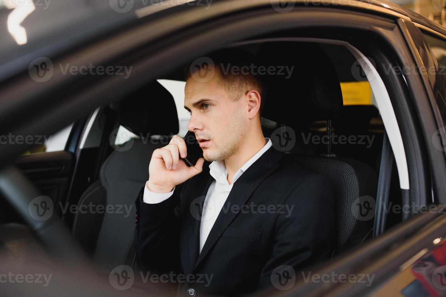 Young businessman sits in luxury car and talks on phone. He looks straight forward. Guy drives car. He holds one hand on steering wheel. It is sunny outside. photo