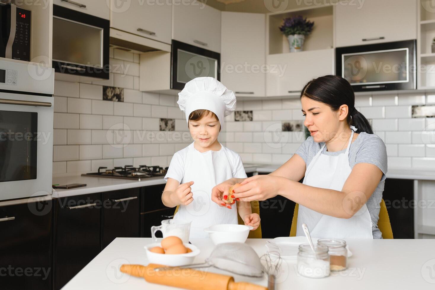 contento madre y su pequeño hijo sacudida crudo huevos en cuenco antes de haciendo masa para hecho en casa Pastelería en el cocina foto