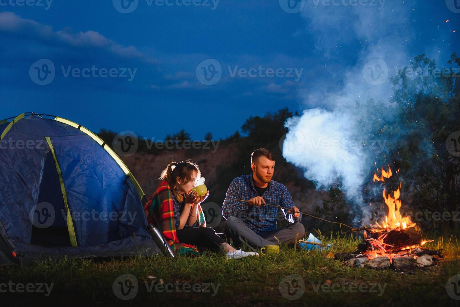 Young couple drinking coffee, and warming up on the campfire in the forest hill in the dusk. The concept of active recreation and travel with a tent photo