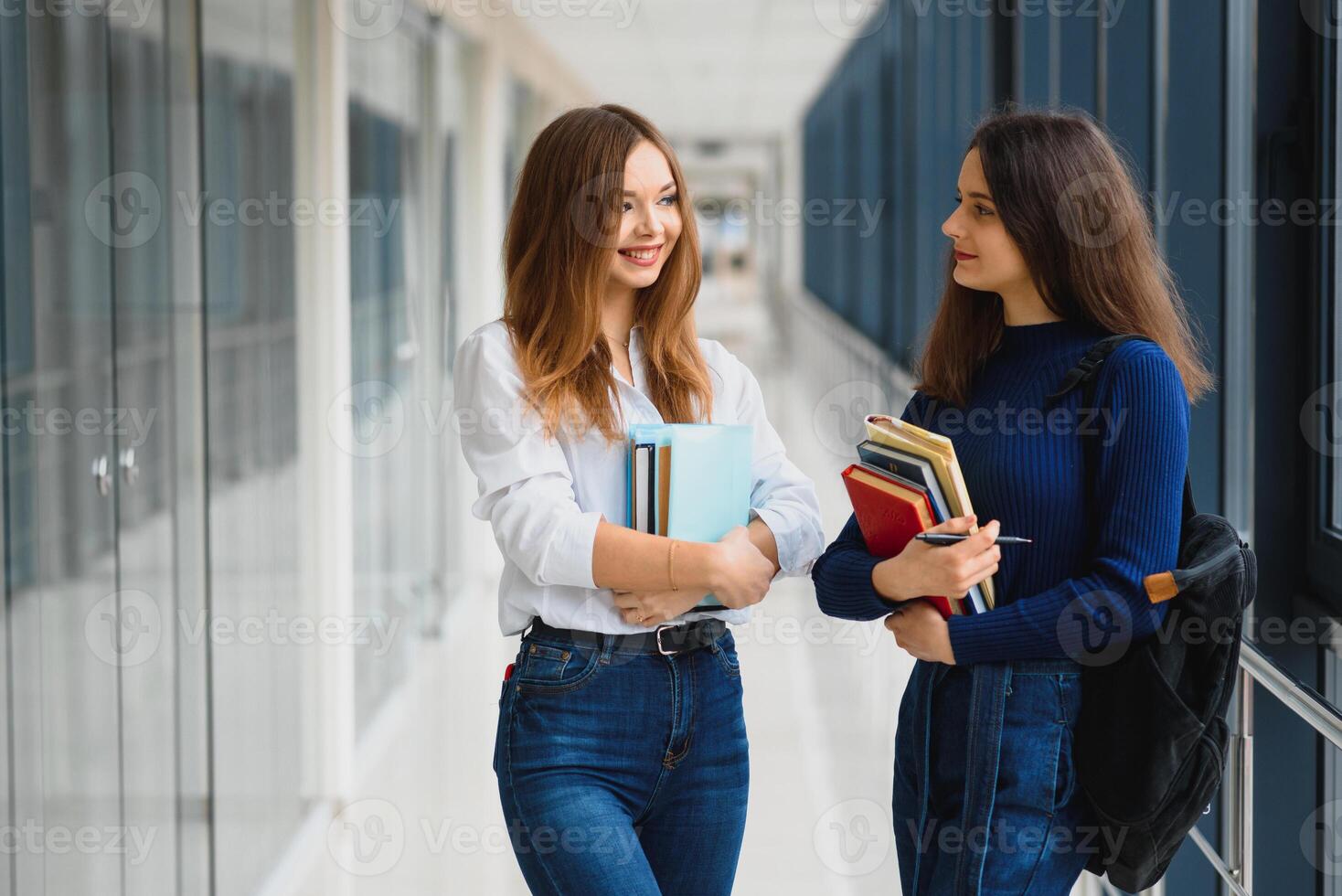 Two young women with book chatting while standing in college corridor. University students in corridor after the lecture. photo