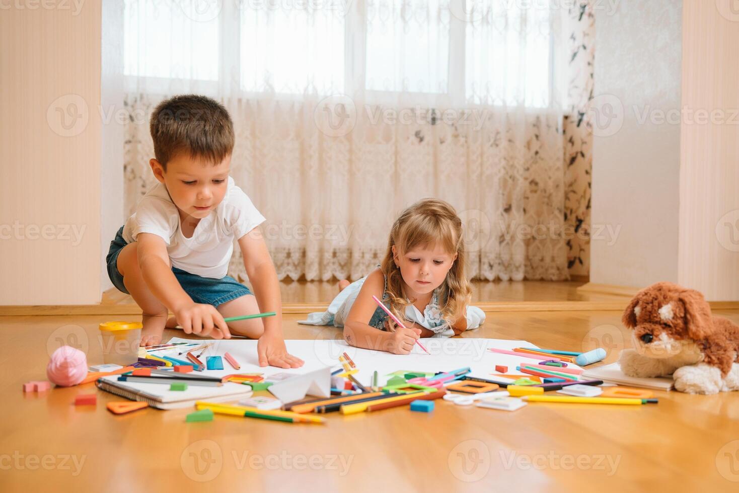 Kids drawing on floor on paper. Preschool boy and girl play on floor with educational toys - blocks, train, railroad, plane. Toys for preschool and kindergarten. Children at home or daycare photo