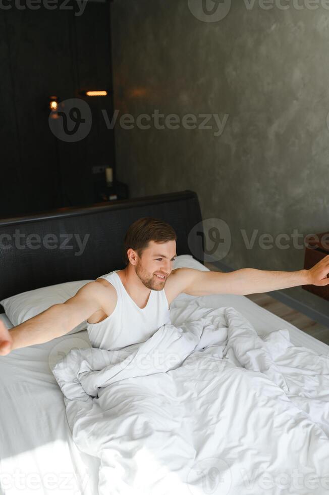 bueno Mañana. joven hombre despertar arriba en cama y extensión su brazos foto