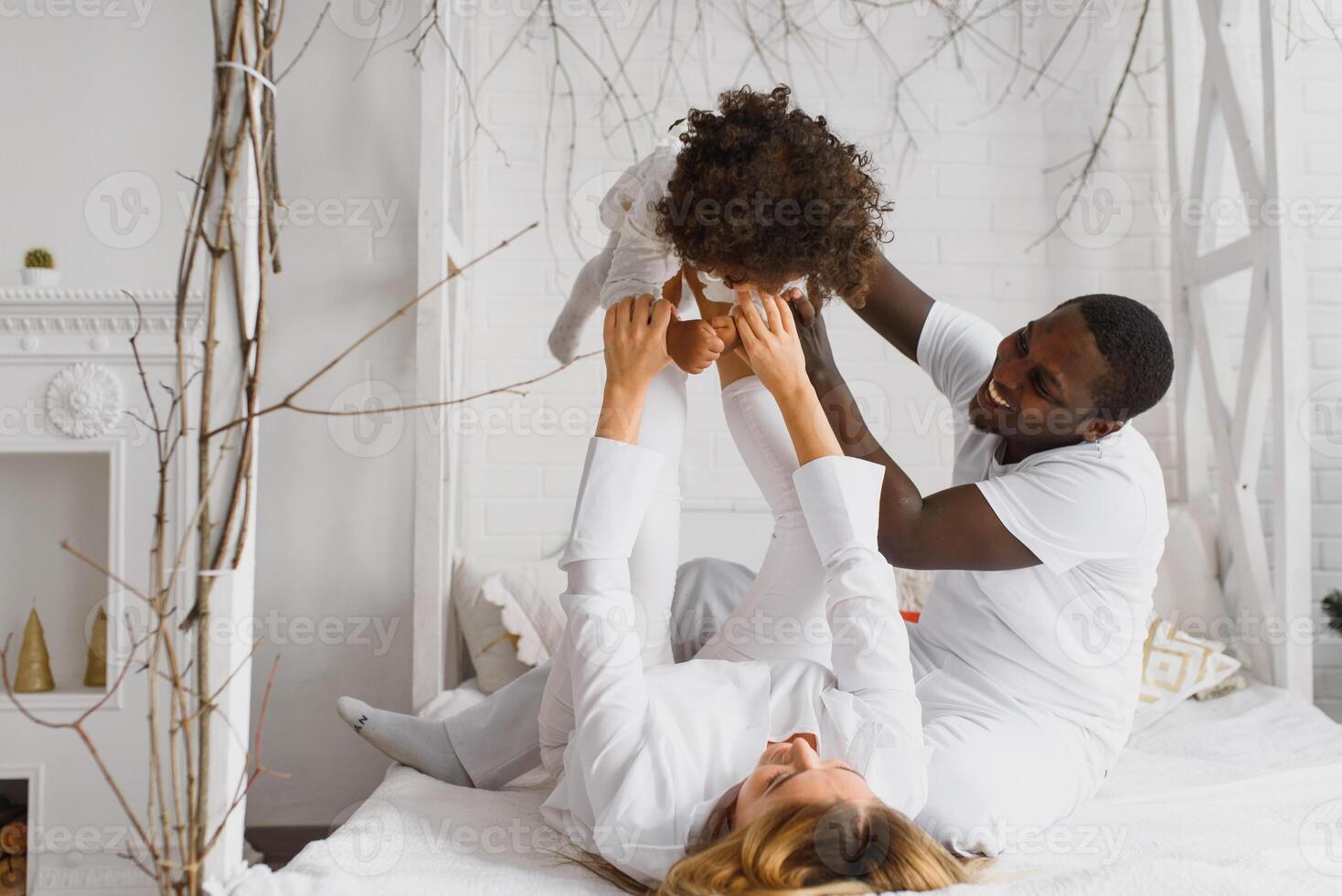 Portrait of happy multiracial young family lying on cozy white bed at home, smiling international mom and dad relaxing with little biracial girl child posing for picture in bedroom photo