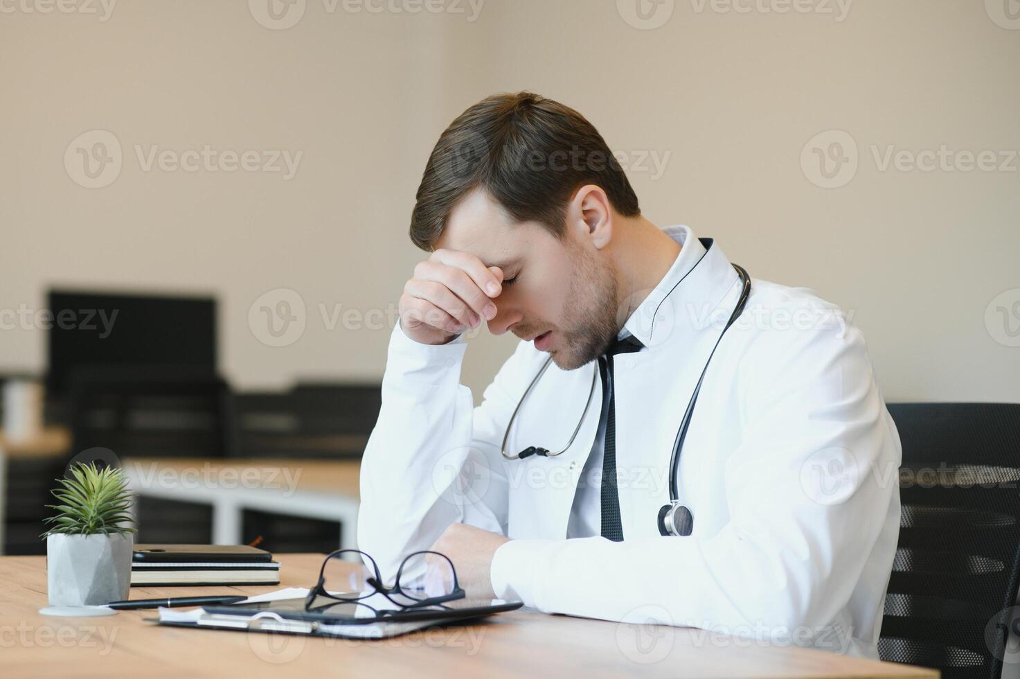 Stressed male doctor sat at his desk. Mid adult male doctor working long hours. Overworked doctor in his office. Not even doctors are exempt from burnout . photo
