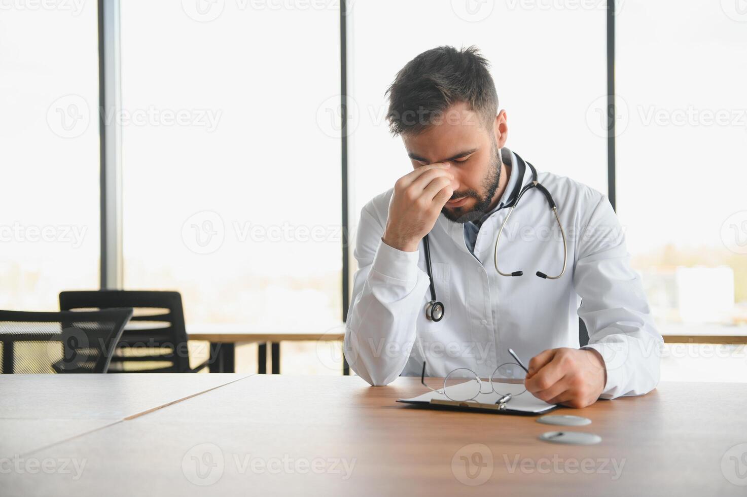Handsome doctor man wearing medical uniform sitting on his workplace tired holding his head feeling fatigue and headache. Stress and frustration concept photo