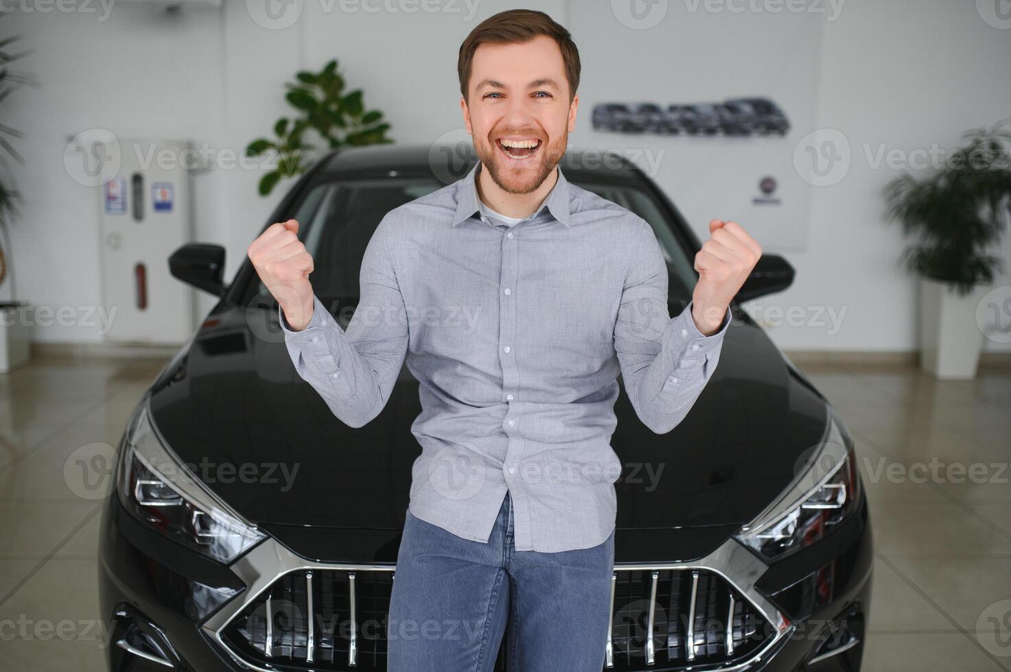 Emotional handsome guy raising hand up and smiling at camera. Rich man buying auto at luxury dealership salon photo