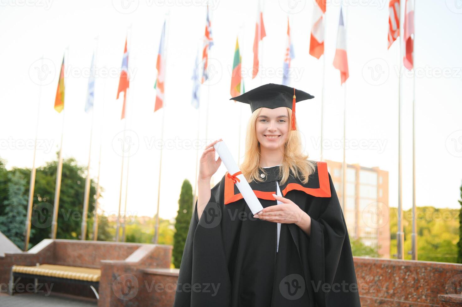 Happy cute caucasian grad girl is smiling. She is in a black mortar board, with red tassel, in gown, with nice brown curly hair, diploma in hand photo