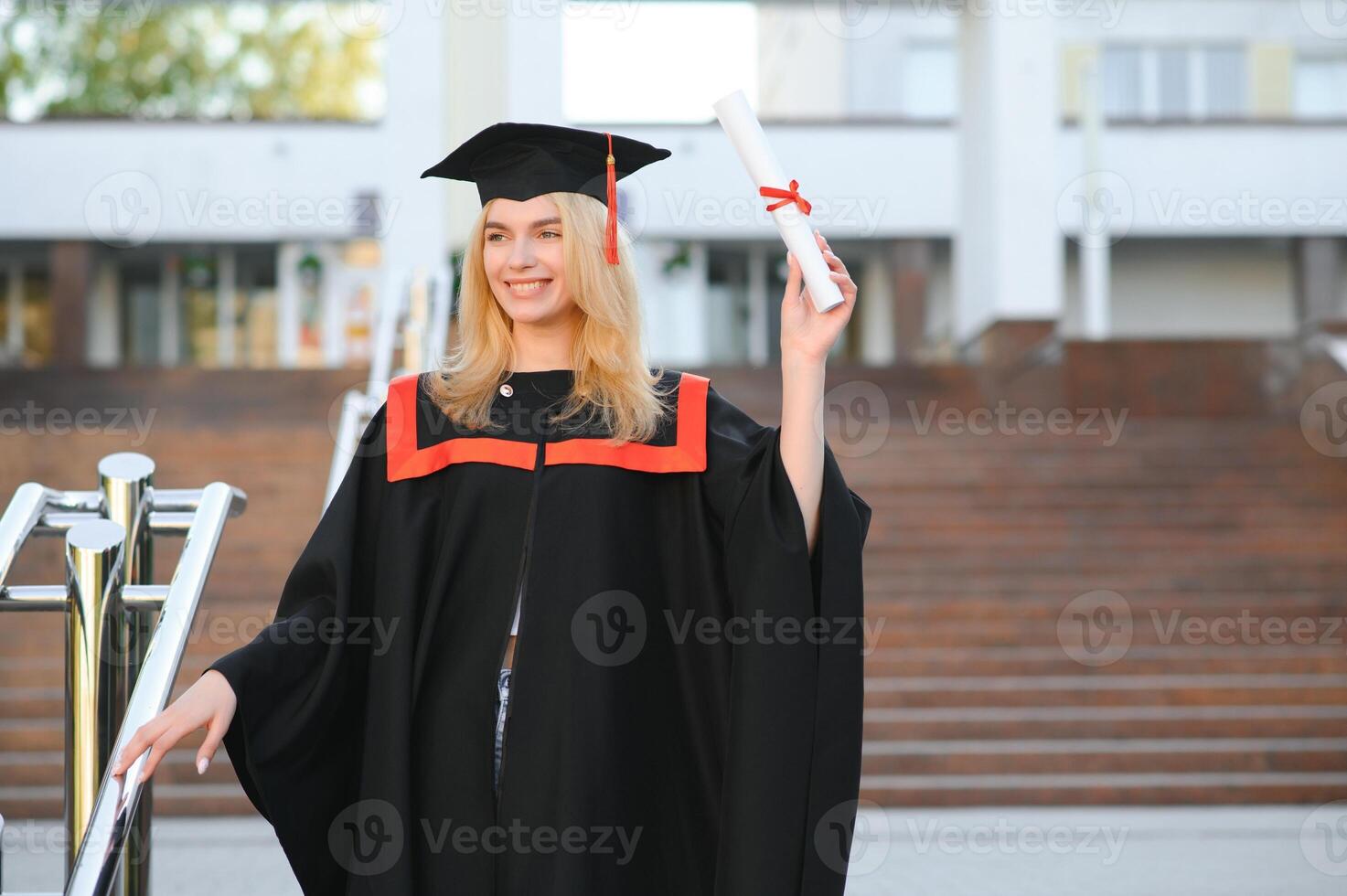 Happy cute caucasian grad girl is smiling. She is in a black mortar board, with red tassel, in gown, with nice brown curly hair, diploma in hand photo