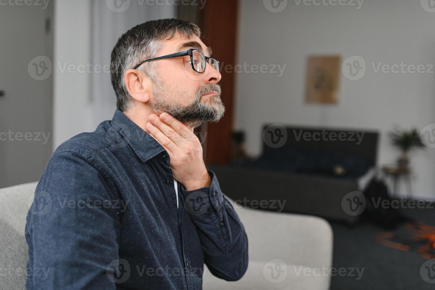 Senior man suffering from cold. Photo of elderly man having sore throat. Elderly Caucasian man touching his throat. Sore throat, tonsillitis or thyroid gland problem. Elder man at home feeling unwell.