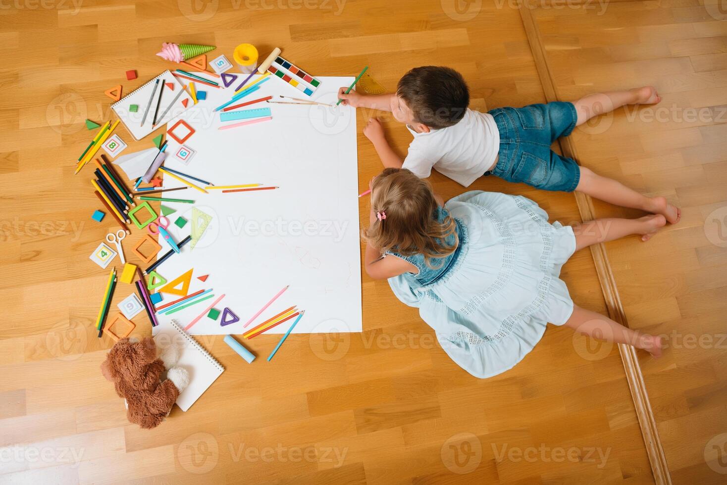 Kids drawing on floor on paper. Preschool boy and girl play on floor with educational toys - blocks, train, railroad, plane. Toys for preschool and kindergarten. Children at home or daycare. Top view. photo