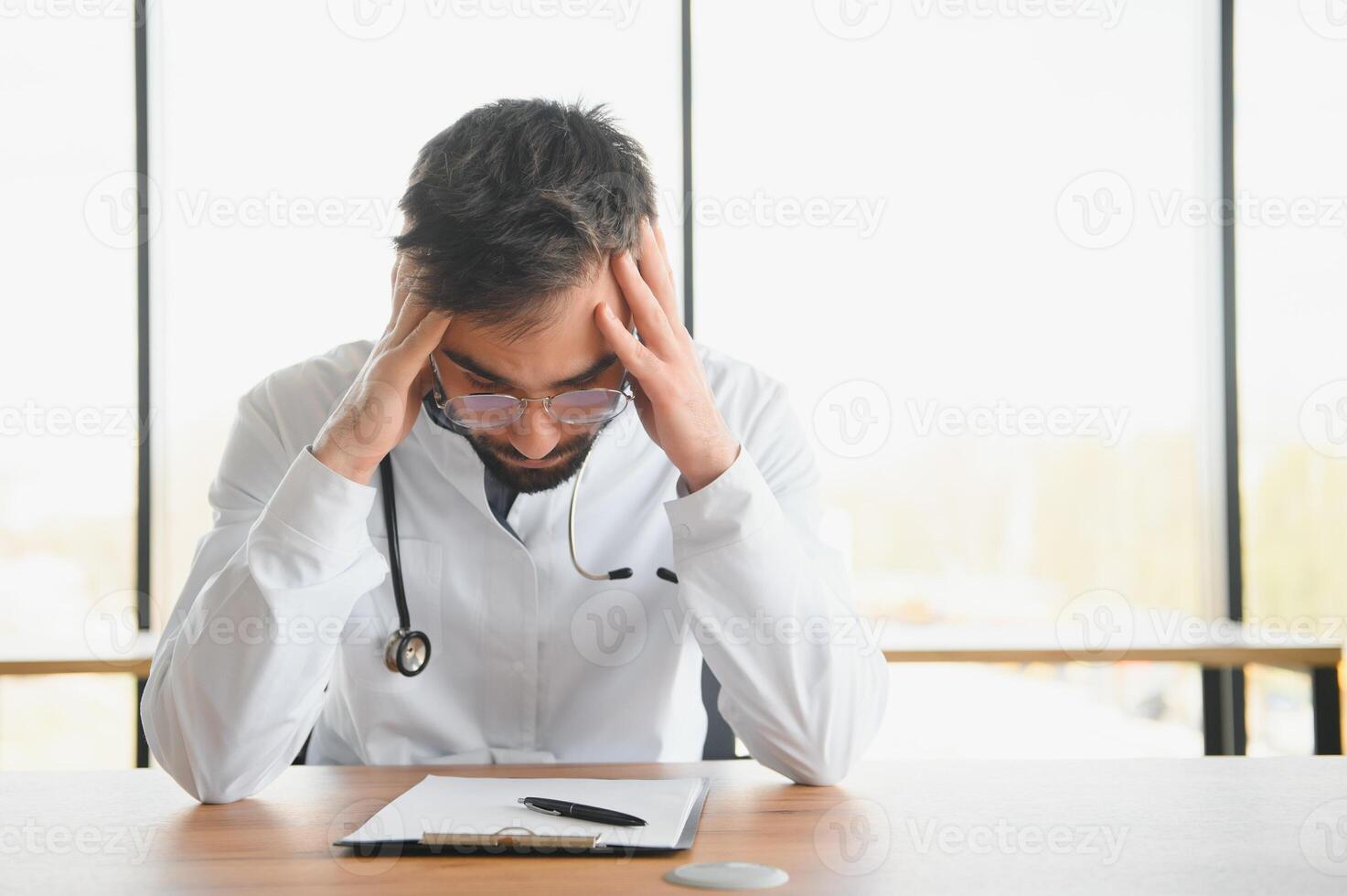 Close-up of upset tired medical worker closing head with hands. Depressed male doctor sitting in office. stethoscope on persons neck. Modern medicine and healthcare concept. photo