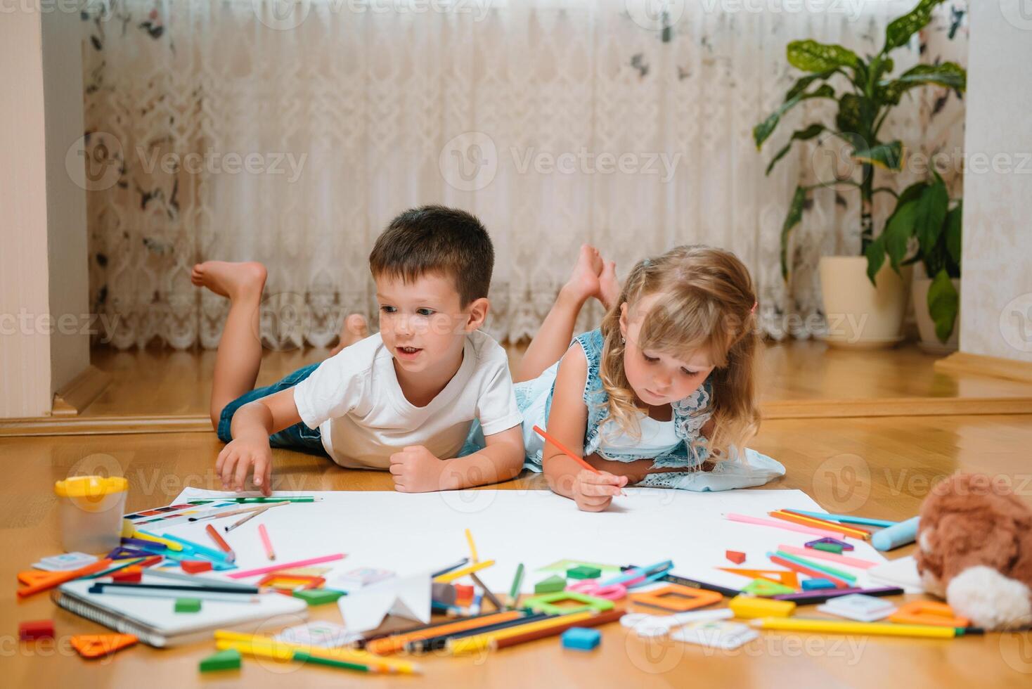 Kids drawing on floor on paper. Preschool boy and girl play on floor with educational toys - blocks, train, railroad, plane. Toys for preschool and kindergarten. Children at home or daycare photo