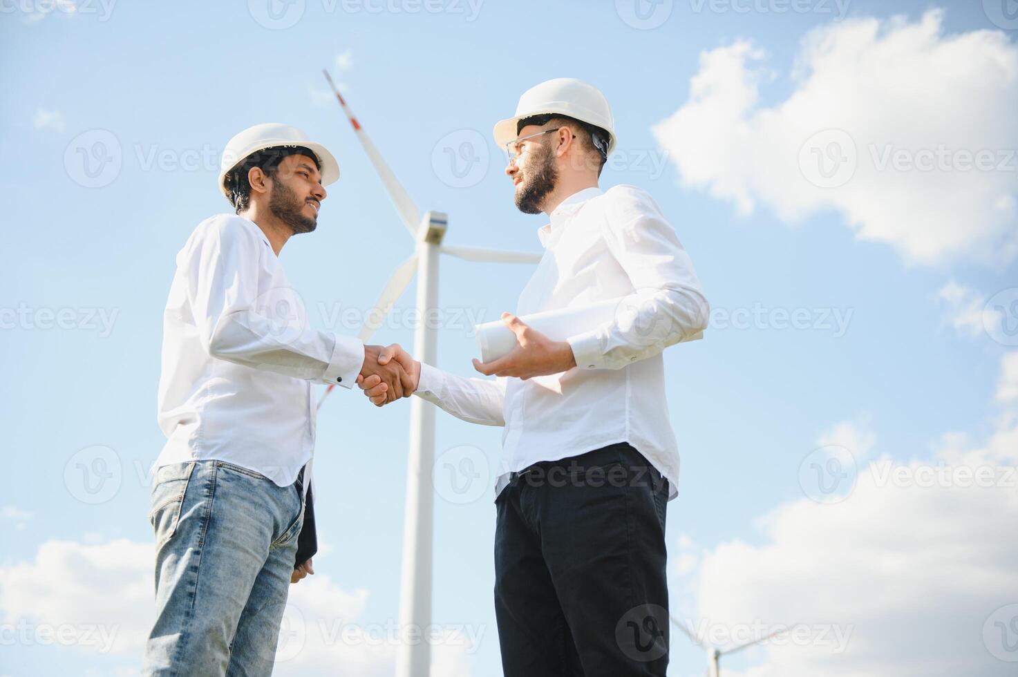 Close up of indian technician and inspector shaking hands while standing on farm with wind turbines. Concept of people, teamwork and eco friendly energy photo