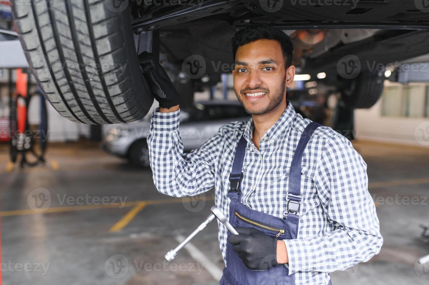 Indian car mechanic standing and working in service station. Car specialists examining the lifted car. Professional repairmen wearing mechanic uniform in blue color. photo