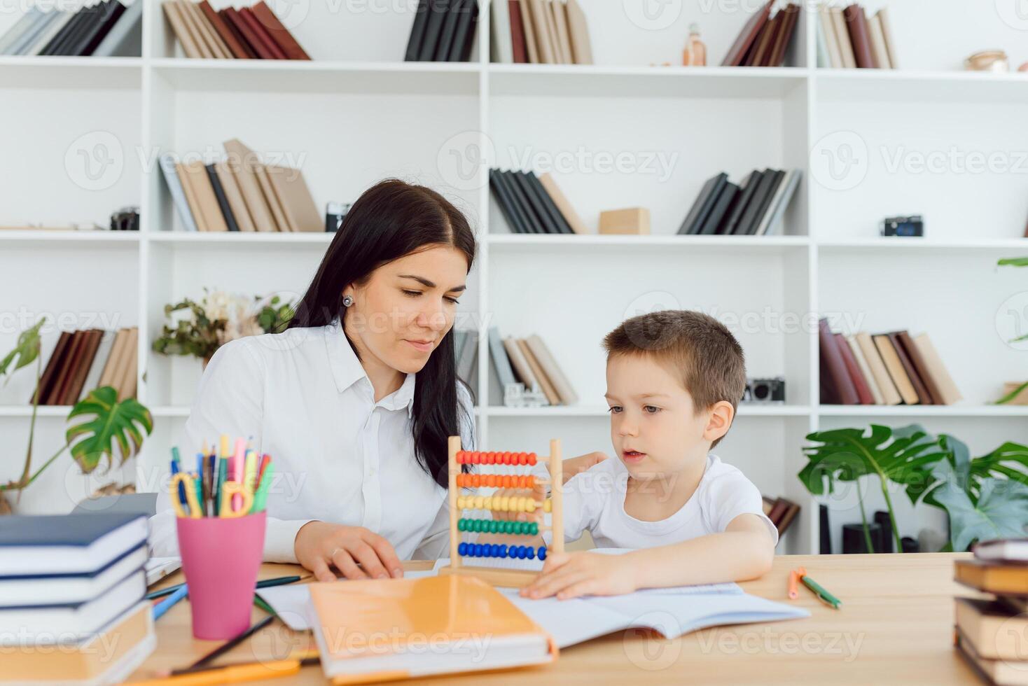 Young female tutor helping little elementary school boy with homework during individual lesson at home photo