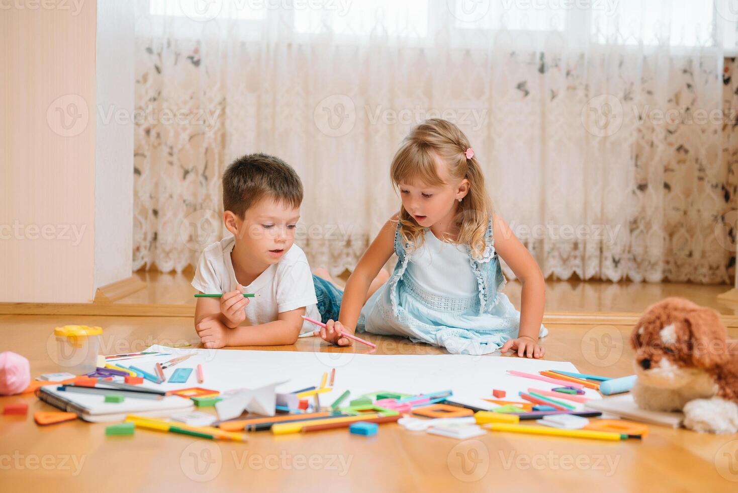 Kids drawing on floor on paper. Preschool boy and girl play on floor with educational toys - blocks, train, railroad, plane. Toys for preschool and kindergarten. Children at home or daycare photo