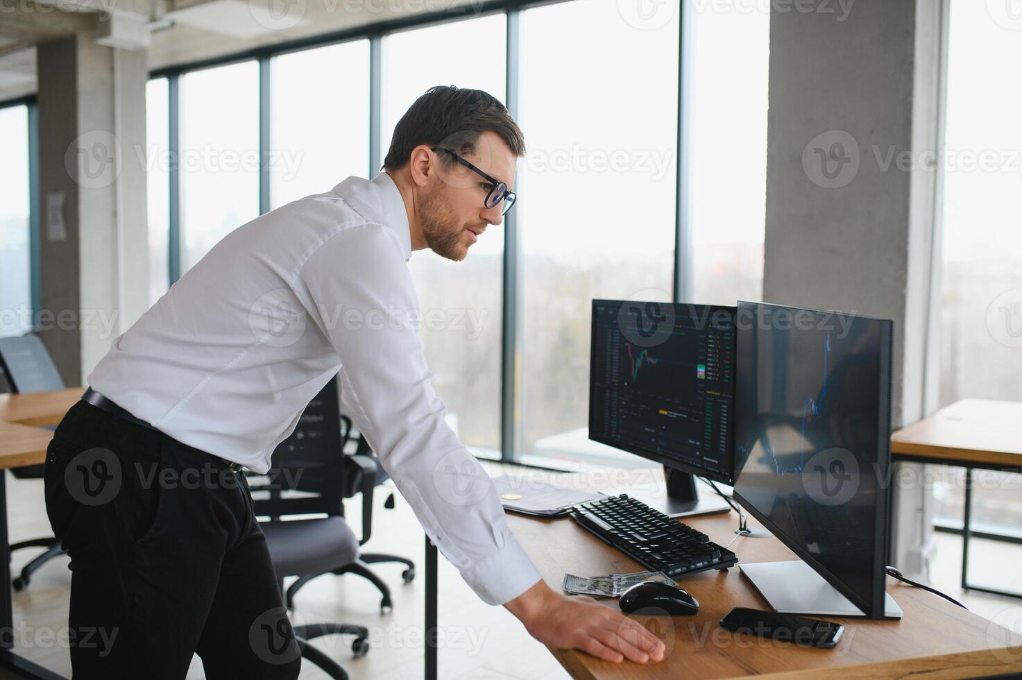 Man trader in formalwear sitting at desk in frot of monitors with charts and data at office browsing laptop checking documents analyzing stocks price changes concentrated. photo