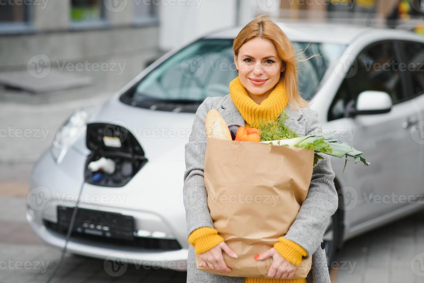 Charging electro car at the electric gas station. Woman standing by the car. Lady with foodstuff photo
