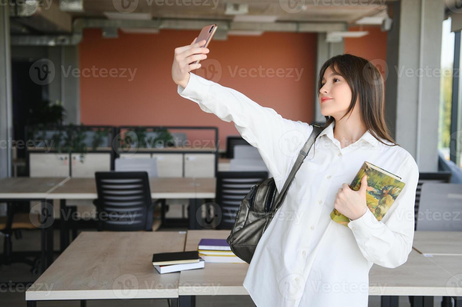 Portrait of a happy cheerful smiling young student schoolgirl lady with long hair standing in empty classroom looking camera take a selfie photo