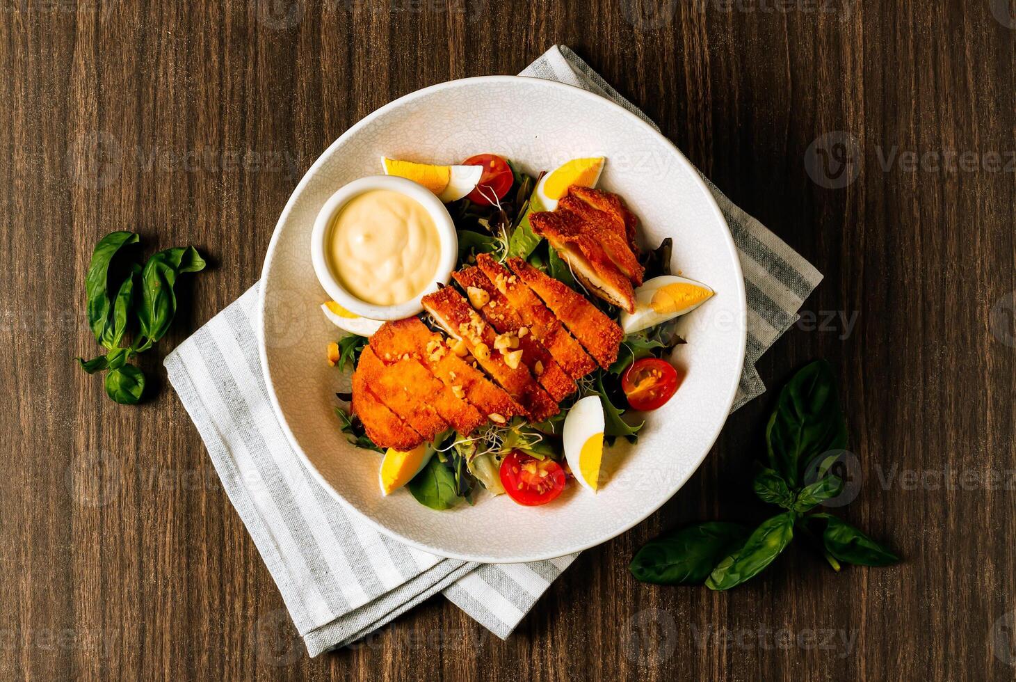 Chicken schnitzel served with fresh salad, boiled egg, tomato and mayo in white bowl on dark wood background top view. photo