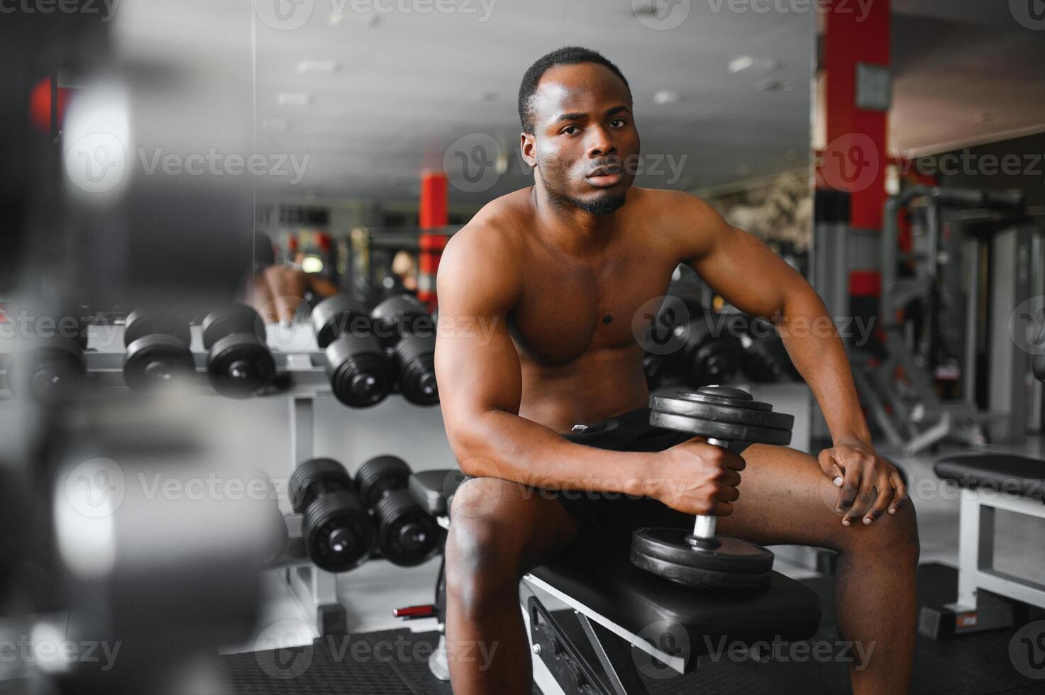 Young African American man sitting and lifting a dumbbell close to the rack at gym. Male weight training person doing a biceps curl in fitness center photo