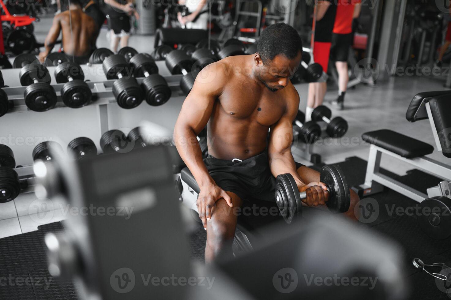 Young African American man sitting and lifting a dumbbell close to the rack at gym. Male weight training person doing a biceps curl in fitness center photo