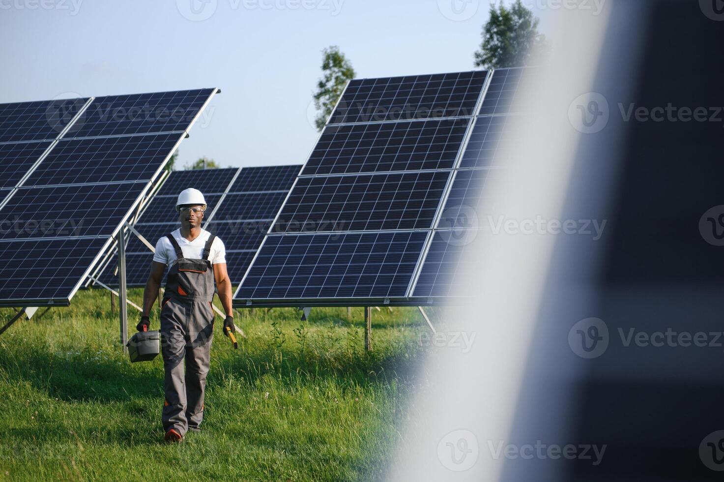 Portrait of young handsome African American craftsman in protective helmet. Man in uniform and with tools standing among solar panels photo